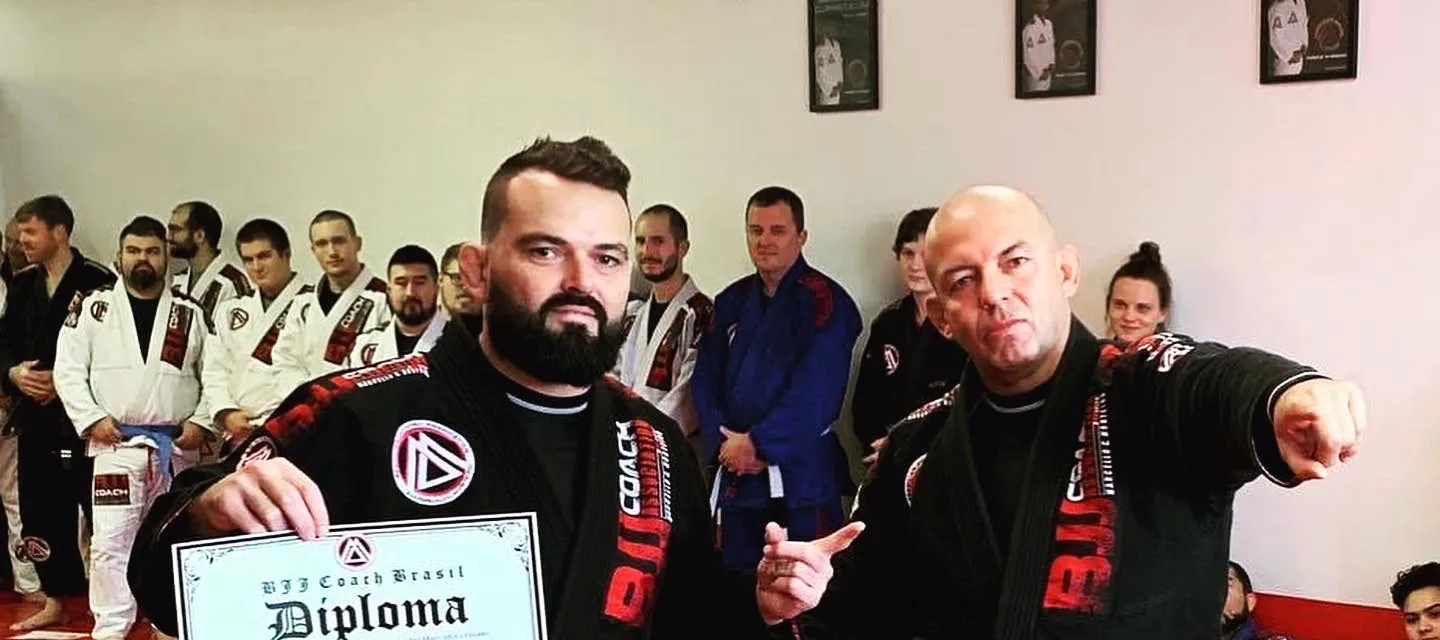 Two adult men in jiu-jitsu gis in the academy, one holding a framed belt diploma