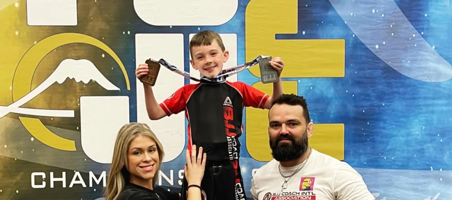 A young boy on a tournament podium holding up two medals, an adult either side of him