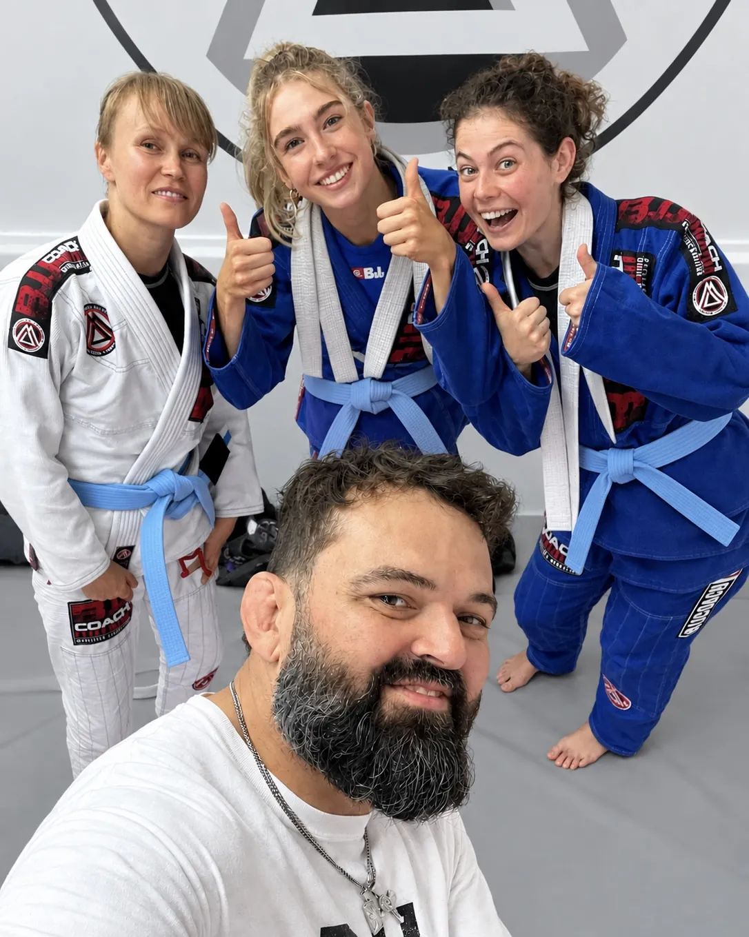 Three students in gi giving thumbs up with their coach in the foreground on the mats